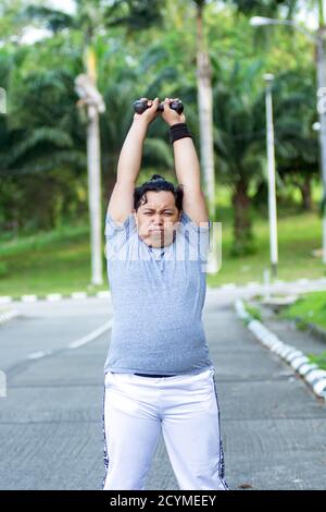 Asian young men with obesity trying to lose weight with barbell exercises and feel not strong in outdoor city parks Stock Photo