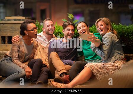Group of diverse friends taking a selfie on a smart phone outdoors at a bar/restaurant, they are celebrating and having fun together. Stock Photo