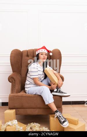 portrait of a young Asian woman wearing a Santa hat and wearing casual clothes sitting on a brown sofa while holding a gift with a Christmas decoratio Stock Photo