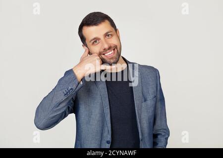 Young businessman man with a beard in a jacket, smiling, making a telephone gesture with his hand and fingers, like talking on the phone. Transfer of concepts. Portrait of a man on a gray background. Stock Photo