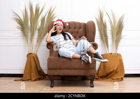 portrait of young Asian woman wearing a Santa hat and wearing casual clothes sitting on a brown sofa indoors Stock Photo