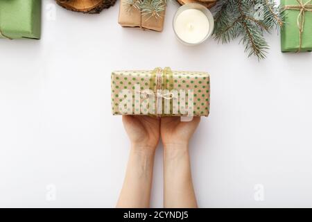 Female hands hold a box with a Christmas gift on white table with Christmas decorations. Christmas, Xmas, winter, new year, zero waste concept. Flat Stock Photo