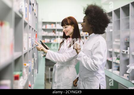Two professional multiethnical pharmacists, African and Caucasian women, checking stock of medicines, using tablet, while standing at the shelve in Stock Photo