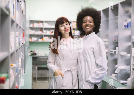 Two happy friendly multiethnic women clinical pharmacists or doctors, posing hugging each other together, smiling to camera in modern hospital Stock Photo