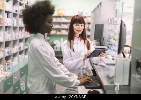 Two young cheerful pharmacists women working together at pharmacy. African woman typing on computer while her Caucasian colleague using tablet. Focus Stock Photo