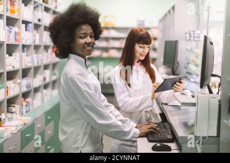 Two young cheerful pharmacists women working together at pharmacy. African woman typing on computer while her Caucasian colleague using tablet. Focus Stock Photo