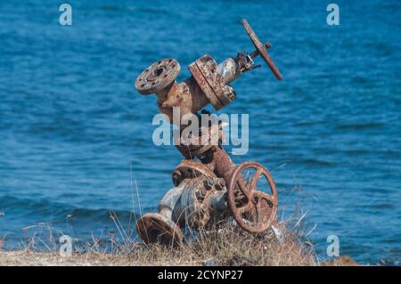 Old rusty weathered aged valve of abandoned pipe system equipment closeup on seashore and blue water background in summer day Stock Photo