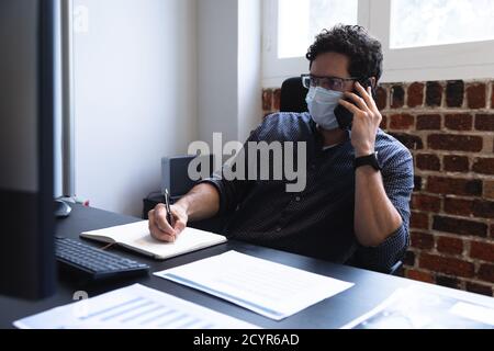 Caucasian man working in a casual office, talking on smartphone, taking notes and wearing face mask. Social distancing in the workplace during Coronav Stock Photo