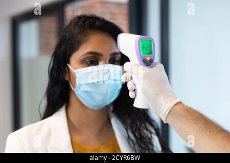Mixed race woman and Caucasian man working in a casual office, wearing face mask, a man taking her temperature. Social distancing in the workplace dur Stock Photo