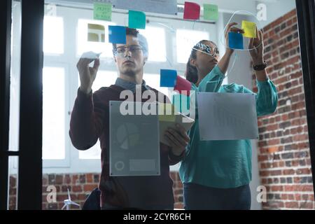 Mixed race woman and Caucasian man working in a casual office, writing on a glass board and talking. Creative business professionals working in a busy Stock Photo