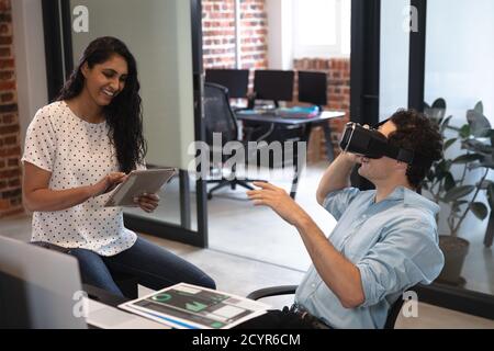 Mixed race woman and Caucasian man working in a casual office, the man wearing vr headset and the woman using a tablet. Creative business professional Stock Photo