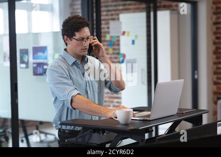 Caucasian man working in a casual office, talking on a smartphone and using a laptop computer. Social distancing in the workplace during Coronavirus C Stock Photo