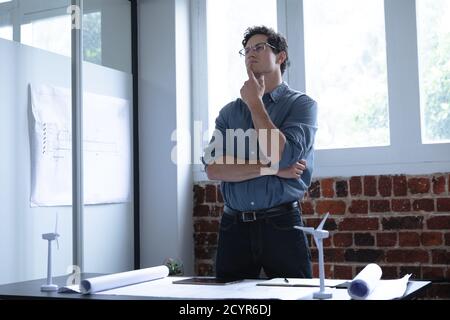 Caucasian man working in a casual office, standing at his desk and thinking. Social distancing in the workplace during Coronavirus Covid 19 pandemic. Stock Photo