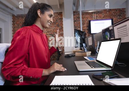 Mixed race woman working in a casual office, sitting at desk, using a laptop computer, waving. Social distancing in the workplace during Coronavirus C Stock Photo