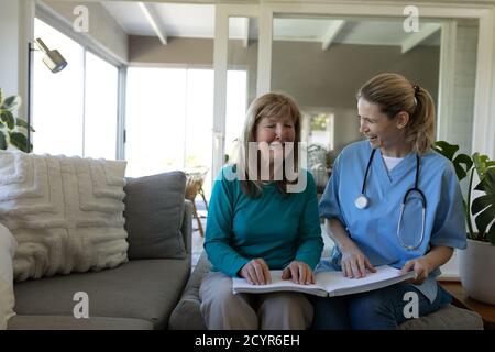 Senior Caucasian woman at home visited by Caucasian female nurse, reading a book using hands. Medical care at home during Covid 19 Coronavirus quarant Stock Photo
