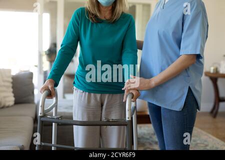 Senior Caucasian woman at home visited by Caucasian female nurse, walking using a walker, wearing face mask. Medical care at home during Covid 19 Coro Stock Photo