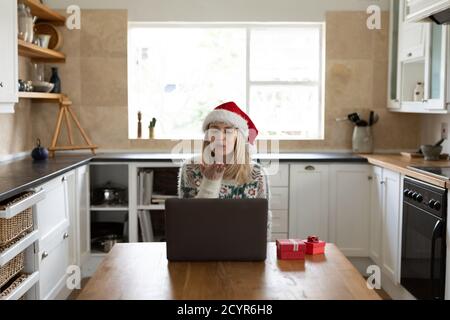 Caucasian woman spending time at home, sitting in kitchen at Christmas wearing Santa hat, using laptop with presents on table. Social distancing durin Stock Photo
