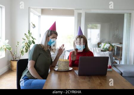 Senior Caucasian woman spending time at home with her adult daughter, sitting in living room with a birthday cake and using laptop. Social distancing Stock Photo