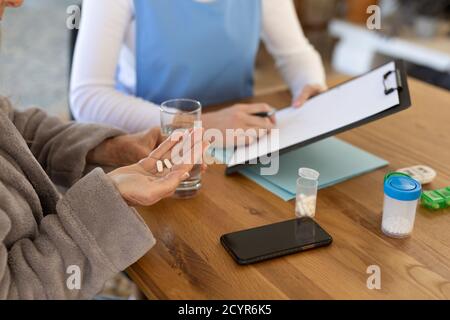 Senior Caucasian woman at home visited by Caucasian female nurse, sitting by table holding a clipboard, woman taking pills. Medical care at home durin Stock Photo