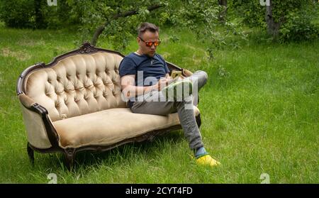 Middle-aged man uses mobile phone resting on light sofa in garden. Sofa is on green grass outdoors. High quality photo Stock Photo