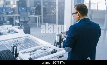 Automotive Design Engineer Looking at Technical Drawing of an Electric Car Chassis Prototype on a Tablet. In Innovation Laboratory Facility Concept Stock Photo