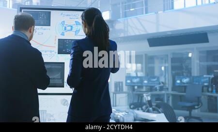 Male and Female Design Engineers are Working and Discussing Something on a Interactive Whiteboard Next to an Electric Car Chassis Prototype. In High Stock Photo