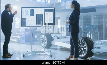 Male and Female Design Engineers are Working and Discussing Something on a Interactive Whiteboard Next to an Electric Car Chassis Prototype. In High Stock Photo