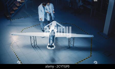 Two Aerospace Engineers Work On Unmanned Aerial Vehicle Drone Prototype. Aviation Scientists in White Coats Talking, Using Tablet Computer. Industrial Stock Photo