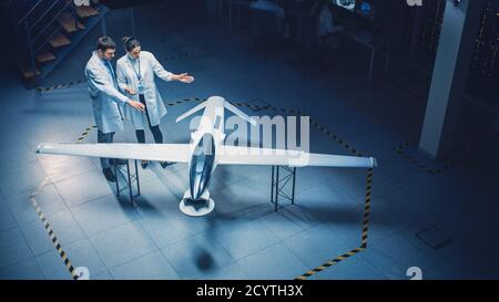 Two Aerospace Engineers Work On Unmanned Aerial Vehicle Drone Prototype. Aviation Scientists in White Coats Talking, Using Tablet Computer. Industrial Stock Photo