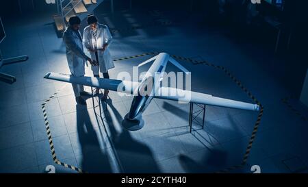 Two Aerospace Engineers Work On Unmanned Aerial Vehicle Drone Prototype. Aviation Scientists in White Coats Talking, Using Tablet Computer. Industrial Stock Photo