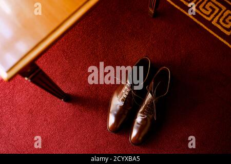 Brown men's shoes with laces on a red carpet in a room by a wooden table. Stock Photo