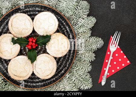 Festive Christmas mince pies on a plate with winter berry holly & icing sugar dusting, snow covered fir & silver fork & napkin on grey grunge  backgro Stock Photo