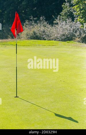 Portrait format photograph of a golf green with a pin and red flag to the left hand side and green copy space to the right. Stock Photo