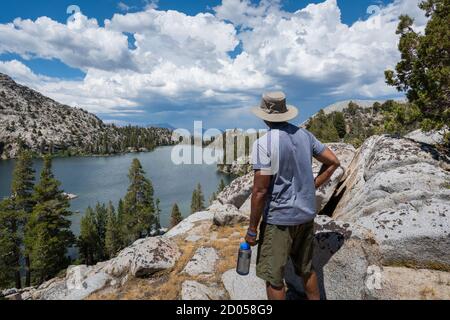 An African American hiker resting at a higher elevation, taking in the views of the lake, trees, mountains and clouds in the distance in California. Stock Photo