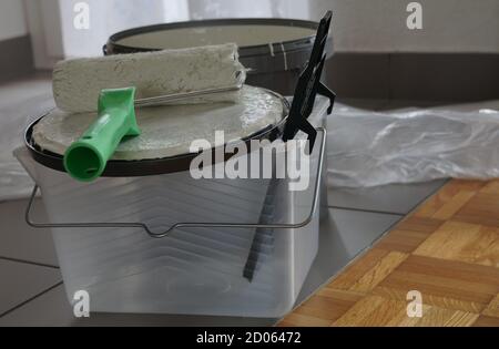 House painting equipment on the floor. There is a paint roller, a bucket of paint and plastic foil to protect the interior from the paint. Stock Photo