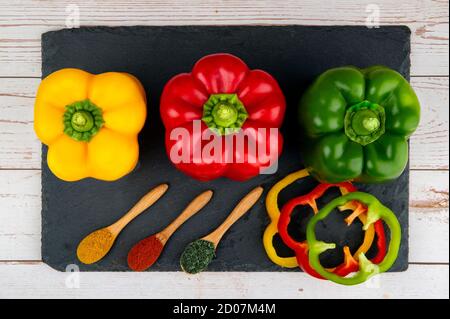 Whole and sliced red, green, yellow peppers and three wooden spoons with spices on black stone table Stock Photo