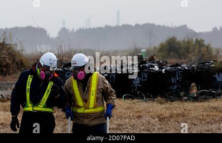 Men wearing radiation protective masks work in front of big black plastic bags containing radiated grass from the decontamination operation as cranes and chimneys of Tokyo Electric Power Co's (TEPCO) tsunami-crippled Fukushima Daiichi nuclear power plant are seen in the background at an area devastated by the March 11, 2011 earthquake and tsunami in Namie town, Fukushima prefecture February 24, 2015. Many residents of Okuma, a village near the stricken Fukushima Daiichi plant, are angry about government plans to dump some 30 million tons of radioactive debris raked up after the March 2011 nucl