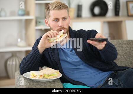 man changing tv channel with remote control while eating sandwich Stock Photo