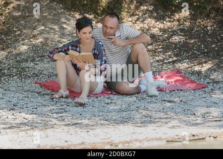 couple looking at book sat on blanket on riverbank Stock Photo
