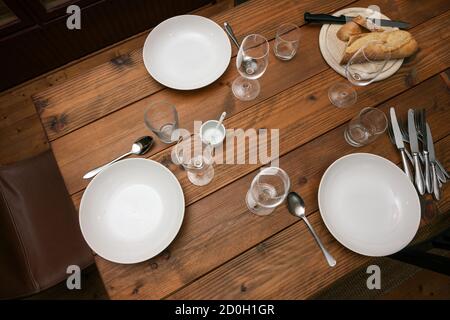 Rustic wooden table half laid with plates, glasses, cutlery and bread for an informal meal with family or friends, selected focus Stock Photo