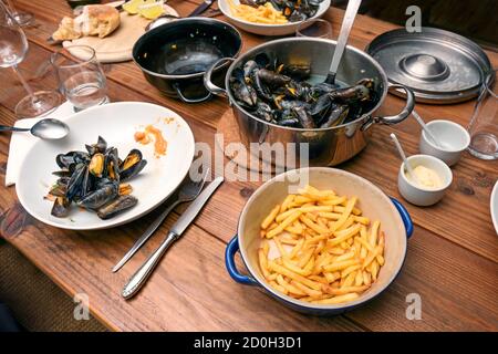 Blue mussels in a pot and French fries, plates, glasses and cutlery on a rustic wooden table, traditional informal meal with family or friends, select Stock Photo