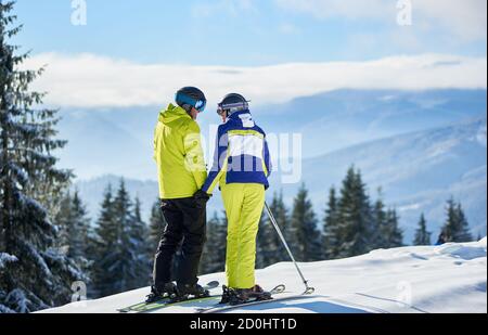 Couple of skiers on mountain edge smiling to each other. Romantic moments of spending time together during winter vacation at ski resort. Mountain panoramic landscape on blurred background. Back view. Stock Photo