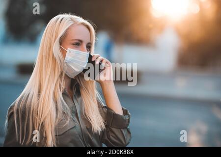 Young blonde woman 24-26 year old wearing medical protective mask talking on phone walking in street outdoors close up. Coronavirus pandemic. Stay at Stock Photo