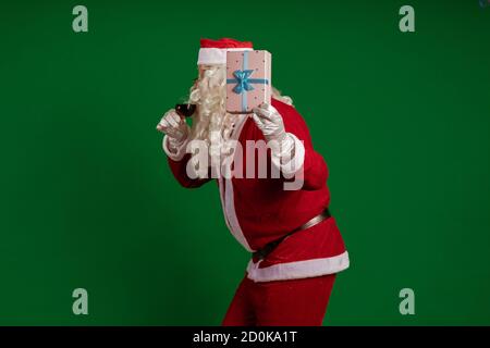 Emotional male actor in a costume of Santa Claus holds one gift box and a glass of wine in his hands and poses on a green chroma background Stock Photo