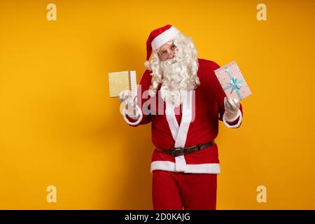 Male actor in a costume of Santa Claus holds two gift boxes in his hands and poses on a yellow background Stock Photo