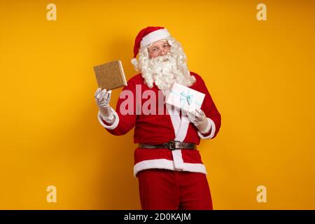 Male actor in a costume of Santa Claus holds two gift boxes in his hands and poses on a yellow background Stock Photo