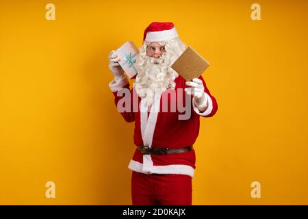 Male actor in a costume of Santa Claus holds two gift boxes in his hands and poses on a yellow background Stock Photo