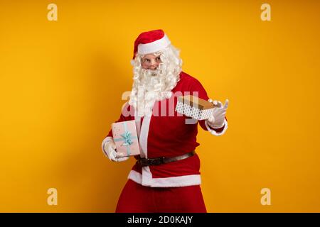 Male actor in a costume of Santa Claus holds two gift boxes in his hands and poses on a yellow background Stock Photo