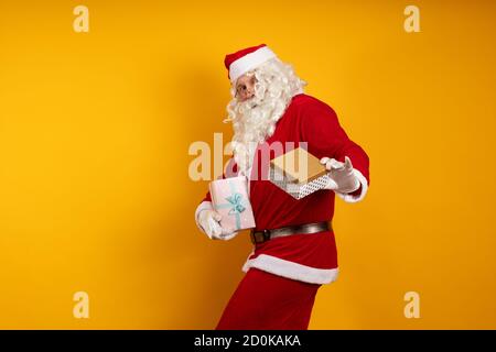 Male actor in a costume of Santa Claus holds two gift boxes in his hands and poses on a yellow background Stock Photo