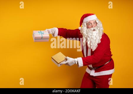 Male actor in a costume of Santa Claus holds two gift boxes in his hands and poses on a yellow background Stock Photo